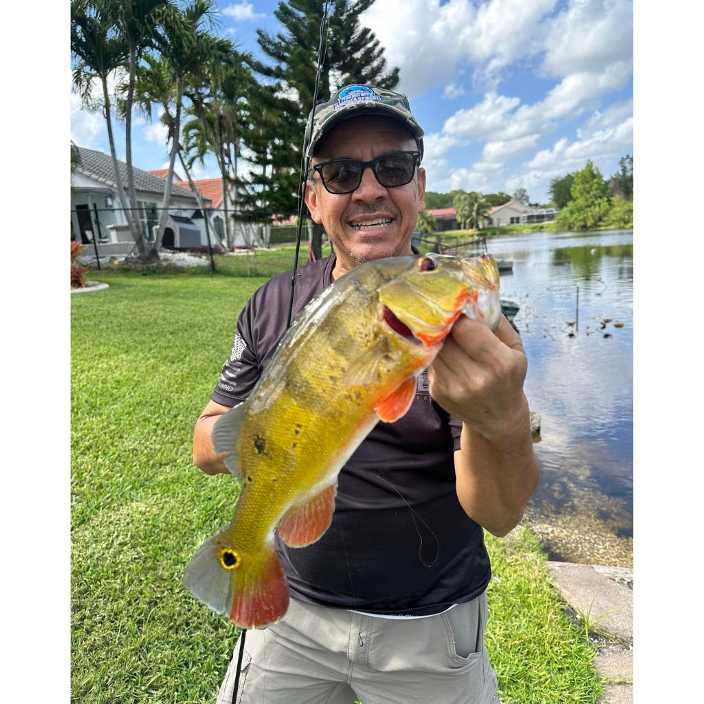 Man holding a large fish by a lake with houses and trees in the background