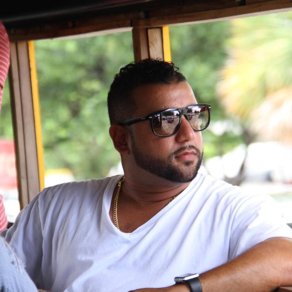 Man wearing sunglasses and a white shirt sitting inside a vehicle with a blurred outdoor background