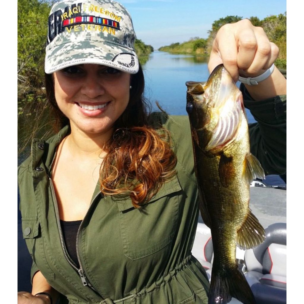 Woman holding a fish by a river, wearing a cap with text and a green jacket.