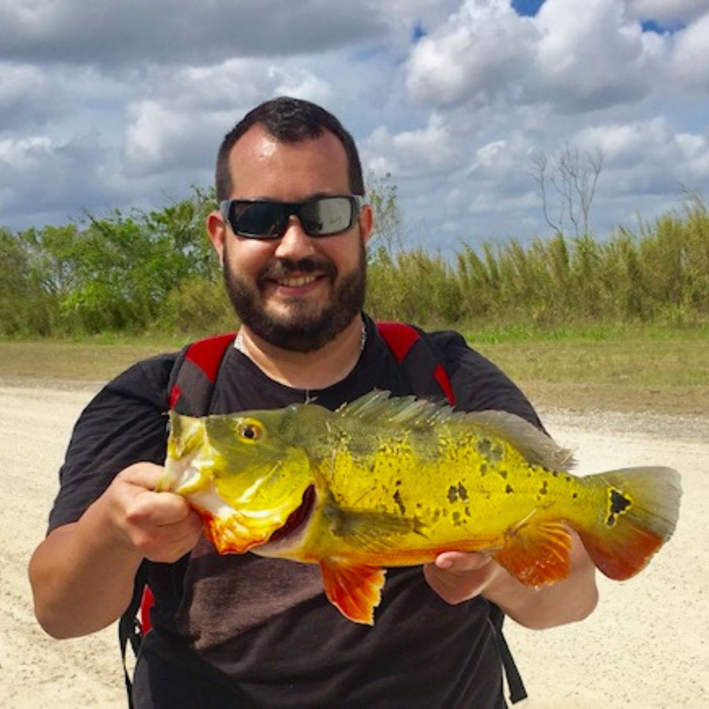 Man holding a large colorful fish outdoors with a cloudy sky and greenery in the background