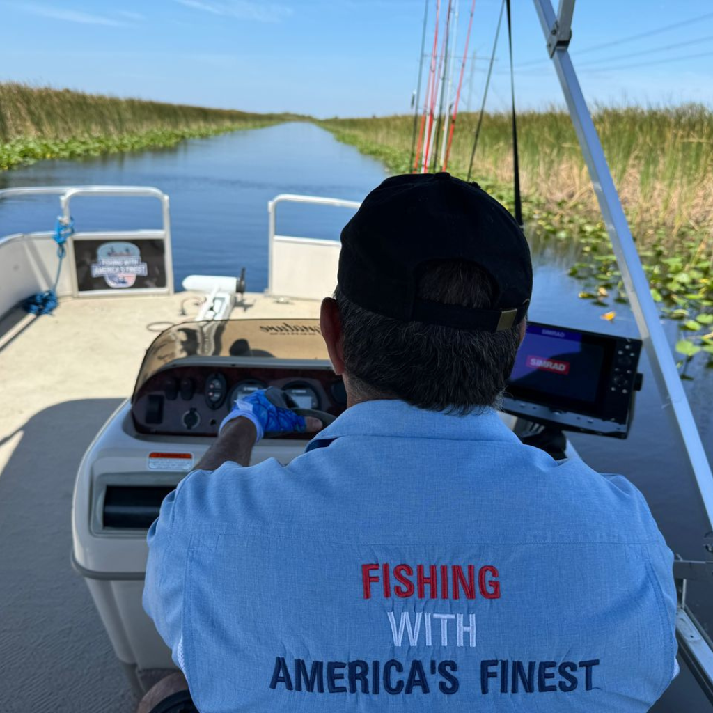 Person on a boat wearing a blue shirt with 'Fishing with America's Finest' text, surrounded by water and greenery.