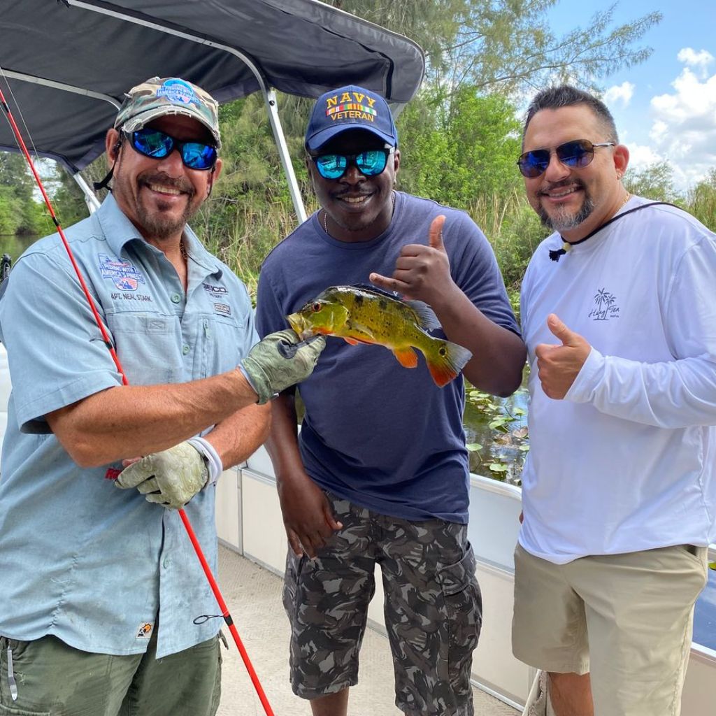 Three men on a boat holding a fish, with sunglasses and casual clothing.